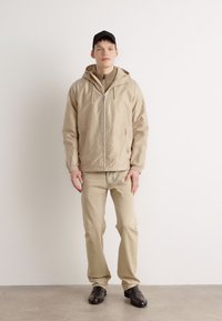 Beige lätt jacka med huva, dragkedjestängning; matchad med beiga byxor och svarta skor. Enkel design, slät textur.