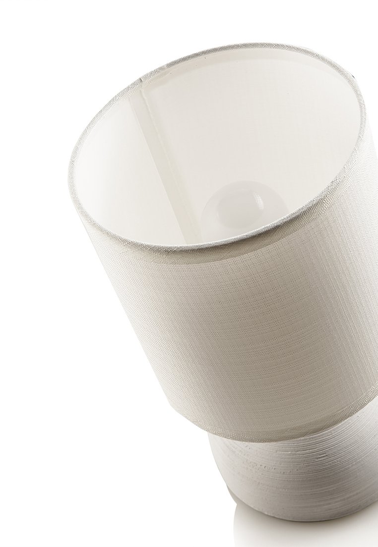 Bílý látkový stínidlo s texturovaným povrchem, umístěné na hladkém, kulatém podstavci. Žárovka viditelná uvnitř stínidla.