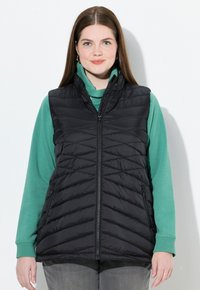 Schwarze, wattierte Weste mit einem Zickzack-Steppmuster, hohem Kragen und mit Reißverschlüssen verschließbaren Taschen, getragen über einem grünen langärmeligen Oberteil.