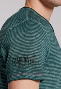 Camiseta de rayas verdes con mangas cortas, escote redondo y un estampado de marca texturizado en negro en la manga izquierda. Detalle de costura visible.