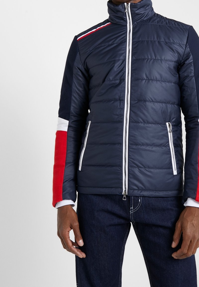 Homme portant une veste matelassée bleu marine avec des fermetures éclair blanches et des manches color block rouge, blanc et bleu, associée à un jean foncé.