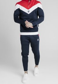 Granatowy dres z czerwono-białym wzorem w zygzak, z pół-zamkiem pod szyją i elastycznym pasem. Posiada akcent z logo oraz zwężane spodnie.