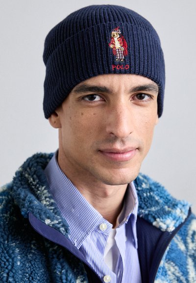 Căciulă tricotată rib de culoare bleumarin, cu un design brodat al unui urs purtând o haină roșie. Materialul are un aspect texturat, subliniind căldura și elasticitatea acestuia.