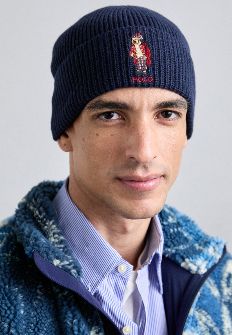 Marinblå ribbad stickad mössa med broderad björndesign som bär en röd kappa. Tyget har en strukturerad yta som betonar dess värme och elasticitet.