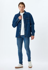 Camisa de denim en azul oscuro, abotonada, con un bolsillo en el pecho. Camiseta blanca de cuello redondo debajo, jeans ajustados azules y zapatillas de color claro.