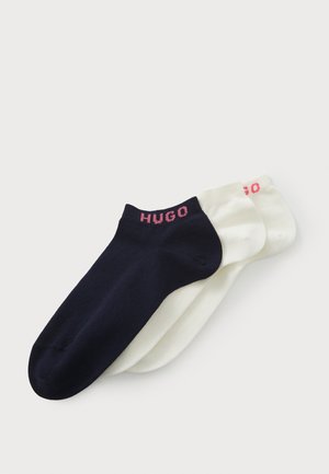 Trois paires de chaussettes courtes : une bleu marine, deux crème. Chacune a un revers côtelé avec "HUGO" en lettres roses. Mélange de coton lisse et léger.