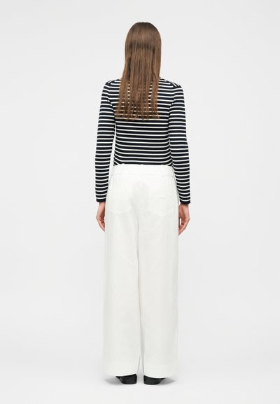 Femme aux longs cheveux bruns portant un haut à manches longues à rayures noires et blanches et un pantalon blanc taille haute à jambes larges, debout de dos.