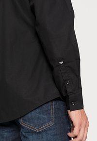 Maglietta nera a maniche lunghe con polsini abbottonati, realizzata in tessuto liscio; dettaglio con hardware grigio; indossata con jeans in denim blu.