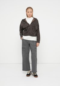 Jaqueta castanha com padrão de leopardo, com fecho, sobre uma camisa branca; combinada com calças cargo cinzas e sapatilhas pretas com detalhes castanhos.