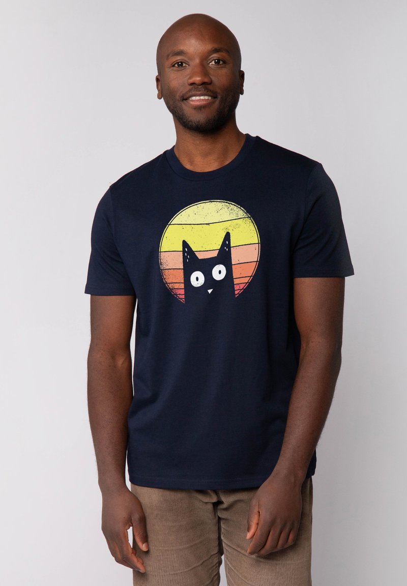 Marineblauwe katoenen T-shirt met een afbeelding van een zwarte kat met grote ogen, geplaatst tegen een cirkelontwerp van een zonsondergang in oranje en gele tinten.