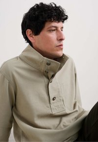 Suéter color caqui claro hecho de suave algodón, con un cuello alto, placket de botones y un bolsillo delantero con detalles de costura.
