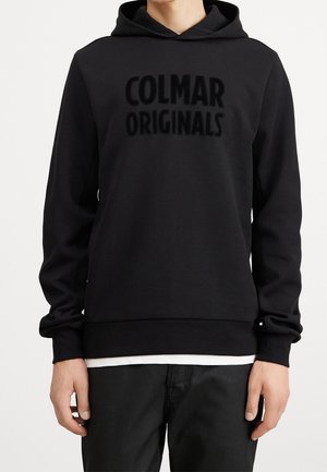 Personne portant un sweat à capuche noir avec "COLMAR ORIGINALS" en lettres noires et grasses, chemise blanche dépassant et pantalon noir, vue de la taille vers le haut.