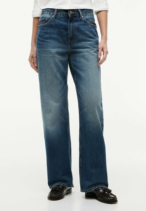 Persoon draagt blauwe rechte spijkerbroek, wit overhemd met opgerolde mouwen en zwarte loafers met kwastjes, staand met ontspannen handen langs het lichaam.