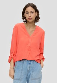 Korallfarbene V-Ausschnitt-Bluse mit langen Ärmeln, die über Knopfdetails und umgeschlagene Manschetten verfügt, kombiniert mit hellblauen High-Waist-Jeans.