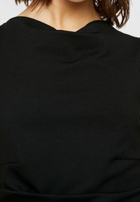 Svart topp med en slät, strukturerad tygkvalitet, klassisk halsringning och subtila sömmar som framhäver en figurnära silhuett.