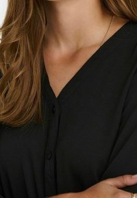 Blusa nera a scollo a V con bottoni frontali, tessuto morbido e maniche con risvolto. Un leggero lucido esalta il design minimalista.