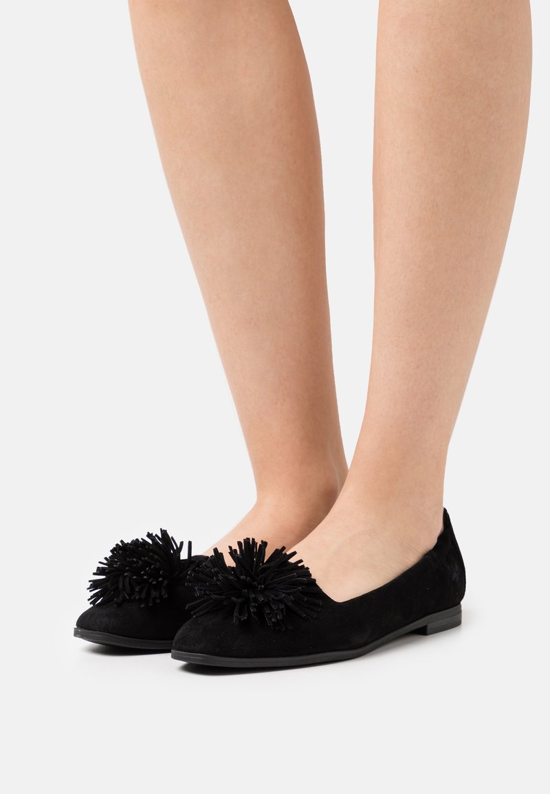 Personne portant des chaussures plates en daim noir avec de grands pompons décoratifs sur les orteils, debout devant un fond clair uni.