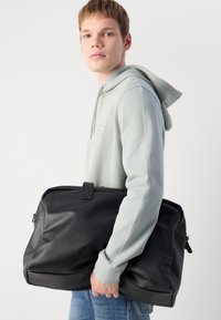 Jonge man in een lichtgrijze hoodie die een grote zwarte leren sporttas vasthoudt, staand tegen een eenvoudige witte achtergrond.