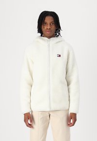 Hvid fleece zip-jakke med hætte, der har et lille broderet logo. Stoffet har en blød, tekstureret overflade.