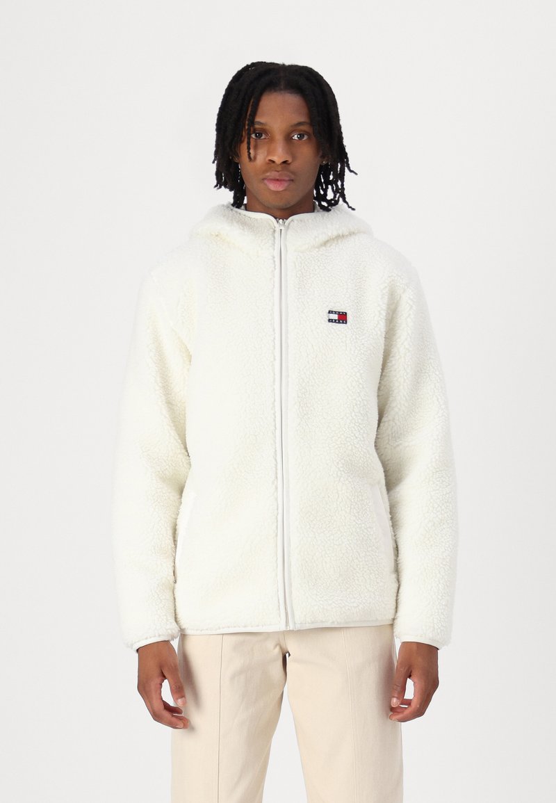 Hvid fleece zip-jakke med hætte, der har et lille broderet logo. Stoffet har en blød, tekstureret overflade.