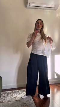 Young woman wearing white shirt and dark wide pants, singing or speaking into a handheld microphone indoors, next to a wall and air conditioner.