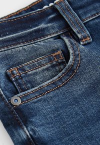 Vaqueros de denim azul con un bolsillo frontal, costuras naranjas y un botón de metal. La textura es suave con una ligera apariencia desgastada.