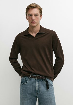Junger Mann mit kurzen blonden Haaren, der ein dunkelbraunes Langarmshirt und hellblaue Jeans vor einem schlichten weißen Hintergrund trägt.