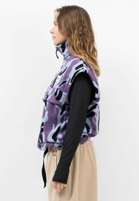 Lila Camouflage-Fleece-Weste mit hohem Kragen, vorderem Taschen-Detail und schwarzem Langarmshirt darunter. Weiche Textur, lockere Passform.