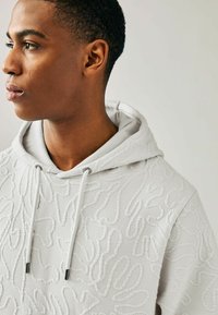 Sudadera de color gris claro con patrones en relieve y texturizados. Cuenta con un cierre con cordón, un interior suave y un corte relajado con capucha.