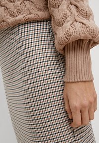 Pull en tricot beige à mailles torsadées avec des poignets côtelés, associé à une jupe à motif pied-de-poule en noir, blanc et beige. Main posée sur la jupe.