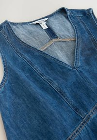 Robe en denim bleu sans manches avec décolleté en V et détails de couture visibles, posée à plat sur une surface blanche.