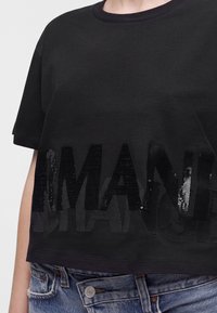 Μαύρο cropped t-shirt με μεγάλο μαύρο κείμενο από παγιέτες μπροστά, φορεμένο με μπλε τζιν παντελόνι.