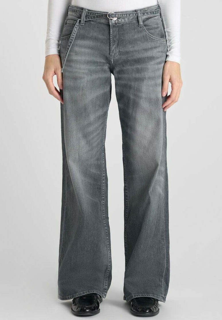 Jean flare en denim gris avec une finition délavée, cinq poches, passants de ceinture et fermeture éclair. Présente des chaussures noires brillantes au niveau de l'ourlet.