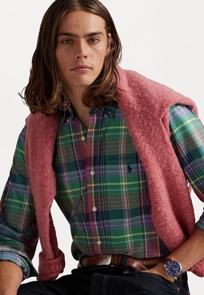 Jeune homme avec des cheveux jusqu'aux épaules portant une chemise à carreaux verts, un pull rose drapé sur les épaules, une ceinture marron et une montre bleue au poignet.