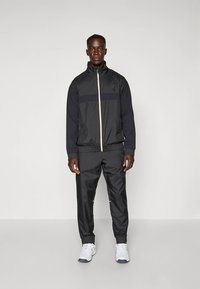 Svart sportdräkt med zipjacka med en kontrasterande blå rand och matchande byxor, tillverkad av slätt, lätt material.