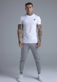 Hvid tætsiddende t-shirt med et lille sort logo; grå joggers med sidelstriber; hvide sneakers. Modellen står med front mod kameraet, armene ned langs siderne.