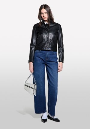 Giacca corta in pelle nera con chiusura a bottoni, jeans a gamba larga blu, calze bianche, scarpe nere a punta e borsa con manico in argento.