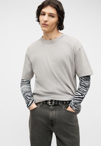 Νεαρό άτομο με μεσαίου μήκους σκούρα μαλλιά που φοράει γκρι κοντομάνικο πουκάμισο πάνω από ασπρόμαυρο μακρυμάνικο με ζέβρα σχέδιο, σκούρο τζιν και μαύρη ζώνη.