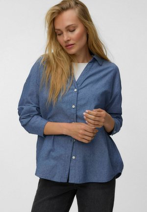 Femme blonde aux yeux fermés portant une chemise bleue ample boutonnée sur un haut blanc et un pantalon noir, debout devant un fond uni.