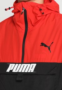 Rode en zwarte windbreaker met een ritssluiting aan de voorkant, verstelbare capuchon, voorzien van een zwart Puma-logo en witte "PUMA" tekst op de onderste sectie.