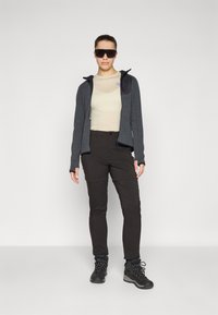 Giacca grigia con zip, shirt beige fitted, pantaloni neri e scarponi da trekking neri con suole testurizzate. Occhiali da sole, vestibilità atletica, design funzionale.