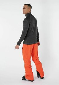 Maglietta grigia a maniche lunghe con colletto alto abbinata a pantaloni da neve arancioni vivaci. I pantaloni hanno una vestibilità comoda e dettagli neri a contrasto sulle caviglie.