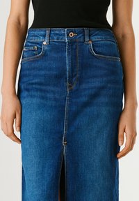 Naine kannab sinist teksaseelikut esilõhikuga, vasevärviliste nööpide ja taskutega, musta varrukateta topiga, käed vabalt külgedel.