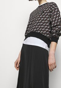 Czarny sweter w wzory z geometrycznym białym motywem, z czarnym ściągaczem u dołu, noszony na białej koszuli, w połączeniu z długą czarną spódnicą.