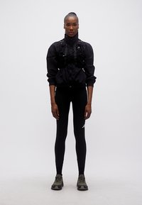 Γυναίκα που στέκεται μπροστά, φοράει μαύρο αθλητικό μπουφάν, κολάν με λευκό λογότυπο και πράσινα αθλητικά παπούτσια σε λευκό φόντο.