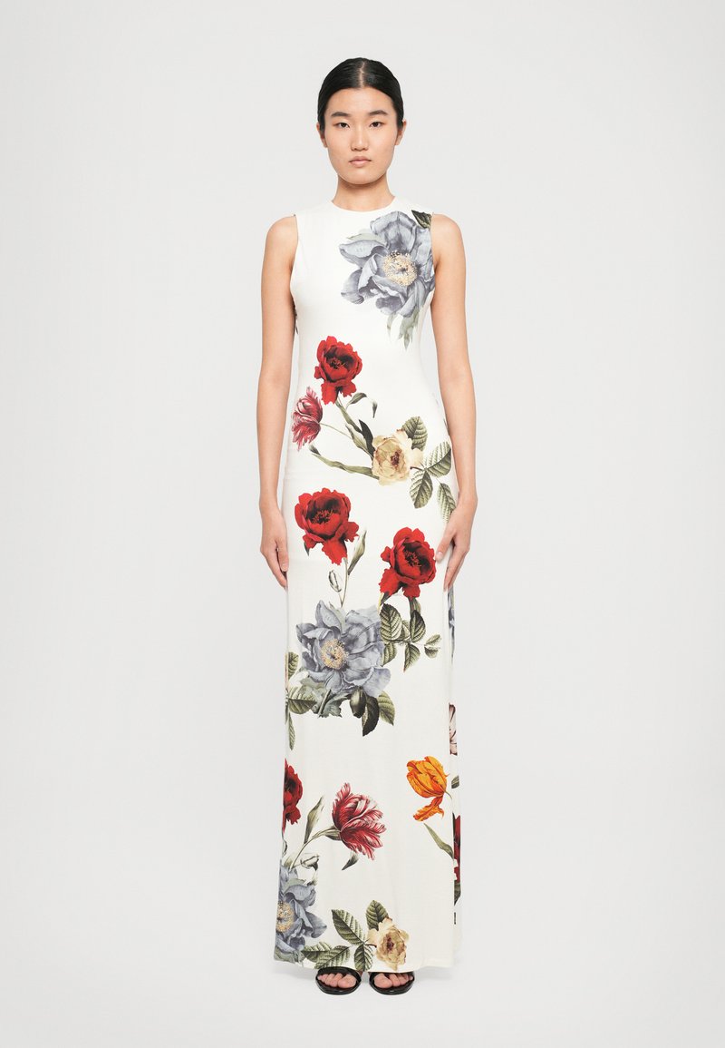 Woman standing straight wearing a sleeveless white maxi dress with large red, orange, and blue floral prints, paired with black sandals.