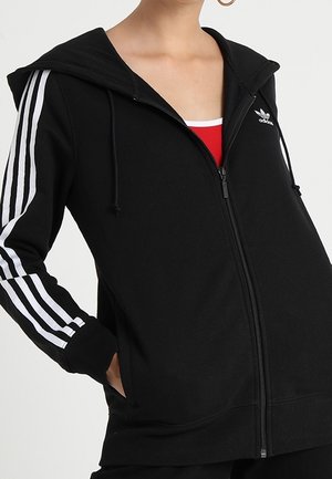Czarna bluza z kapturem na zamek marki Adidas z białymi paskami na rękawach i logo na piersi, noszona na czerwono-białej bluzce.