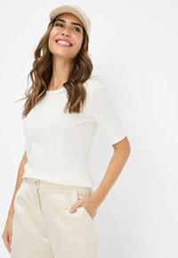 T-shirt bianco a righe con maniche corte e scollatura rotonda, abbinato a pantaloni beige chiaro. Il modello indossa un berretto beige.
