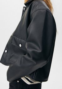 Personne portant une veste en cuir noire à boutons-pression argentés et un pantalon noir, debout devant un fond clair uni.