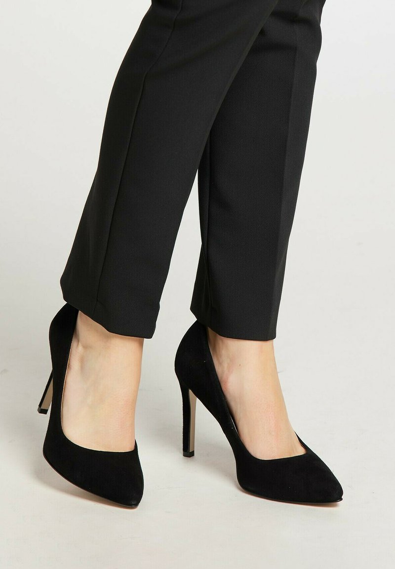 Pantofi cu toc înalt din suede negru, cu vârf ascuțit și toc subțire, asortați cu pantaloni negri mulați, evidențiind un design elegant.